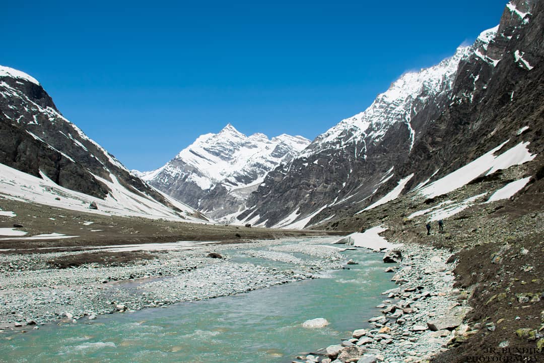 Gupt Khal trek, one of the most difficult treks in India, featuring a wide river, snow-covered hills, and majestic mountain scenery.