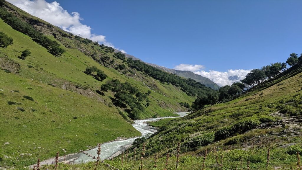River flowing through green fields under a clear blue sky on the Lamkhaga Pass Trek, one of the most difficult and peaceful treks in India