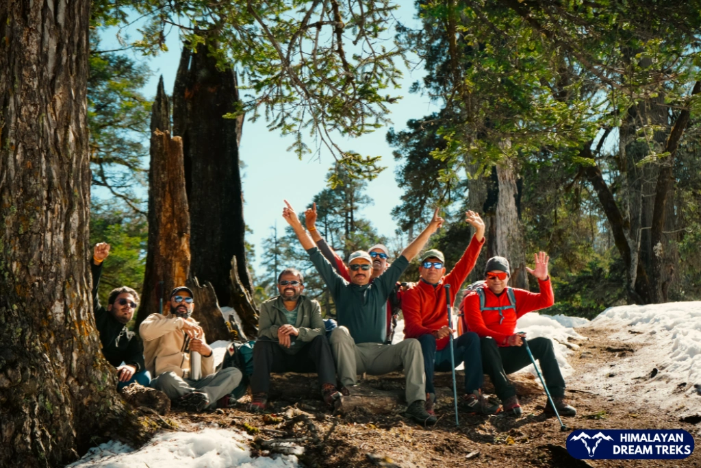 Group of hikers resting beside snow-covered paths and pine trees on the Kuari Pass Trek